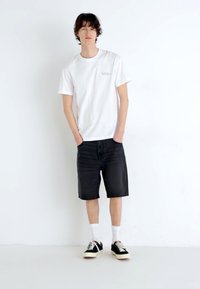 Weißes T-Shirt mit kleinem aufgedrucktem Logo, schwarze knielange Shorts, weiße Socken und schwarze Sneaker mit cremefarbenen Sohlen auf einem hellen Hintergrund.