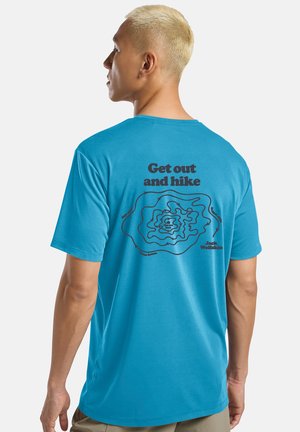 Mann mit kurzem blondem Haar, der ein blaues T-Shirt trägt und eine Wander-Topografiekarte zeigt; auf der Rückseite steht der Text "Rausgehen und wandern".