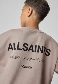 Gaišpelēka pārāk liela T-krekla, kuram aizmugurē ir izteikta melna "ALLSAINTS" uzraksts, ar papildu tekstu japāņu valodā un "LONDON" zem tā.