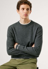 Suéter gris oscuro con cuello redondo y mangas largas, que presenta un cuello interior blanco en contraste y un detalle de logo en el puño.