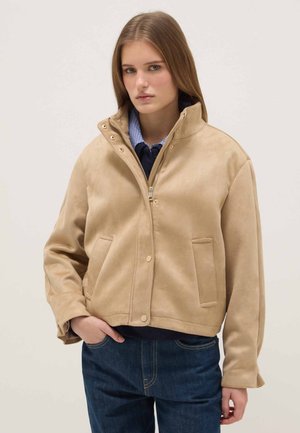 Joven mujer con chaqueta beige con cremallera sobre una camisa azul marino y jeans azules, de pie con una mano en el bolsillo, sobre un fondo liso.