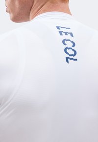 Nahaufnahme des oberen Rückens und Nackens einer Person, die ein weißes Hemd mit blauem vertikalem Schriftzug „LE COL“ nahe der Schulter trägt.