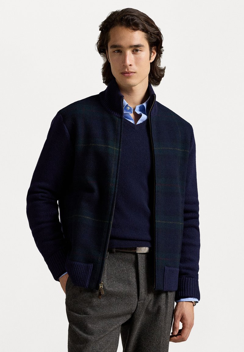 Navy zip-up jacket with textured knit fabric, featuring green and yellow plaid accents, high collar, and ribbed cuffs. Worn over a blue shirt.