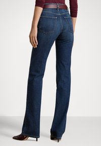 Γυναίκα που φοράει σκουρομπλε τζιν παντελόνι με ίσια γραμμή, μπορντό τοπ και καφέ παπούτσια με τακούνι, εμφανίζεται από πίσω σε απλό φόντο.