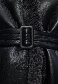Îmbrăcăminte din piele neagră cu un guler moale, texturat din blană de miel de culoare gri închis și o curea neagră rezistentă cu o cataramă rectangulară.