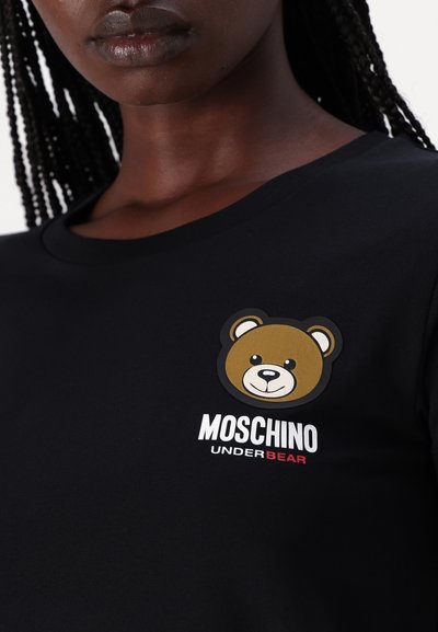 Juodas medvilnės marškinėliai su lokio grafikos uždėklu ir tekstu "MOSCHINO UNDERBEAR" baltos ir raudonos spalvos. Trumpomis rankovėmis ir apvaliu iškirpimu.