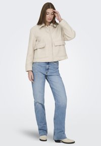 Ung kvinne med langt brunt hår iført en kremfarget jakke med store lommer, lyseblå rette jeans og kremfargede støvler, står med hånden på pannen.