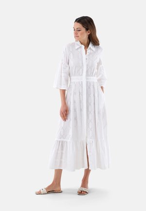 Weißes mittellanges Kleid mit einem Knopfkragen, bestickten geometrischen Mustern, ausgestellten Ärmeln und einem Rüschensaum. Getragen mit geflochtenen Sandalen.