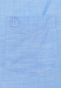 Helles blaues Stoff mit einer subtilen Textur, das eine Brusttasche mit abgerundetem Saum und ein gesticktes Logo in der oberen linken Ecke aufweist.