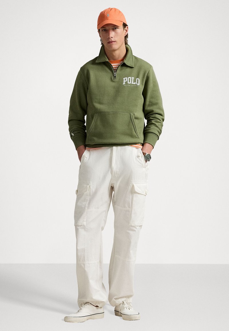 Groene fleece kwart-rits pullover met "POLO" logo, witte cargo broek en een oranje pet. Outfit omvat een gestreept shirt eronder.