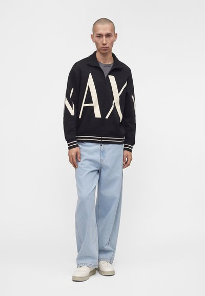 Homme portant une veste noire zippée avec de grandes lettres "NAX", un jean large bleu clair, des baskets blanches, debout devant un fond blanc uni.