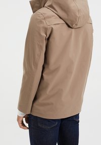 Giacca impermeabile beige con cappuccio, caratterizzata da una texture liscia e un design minimalista. Abbinate a jeans blu scuro, viste da dietro.