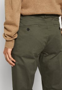 Olivgröna byxor med en slät textur, som har två bakfickor med svarta knappar. Midjan sitter vid den naturliga midjan.