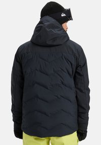 Man in een zwarte gebreide muts, zwarte gewatteerde hoodie, skibril, zwarte handschoenen en felgele broek, met zijn rug naar de camera op een witte achtergrond.