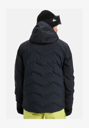 Muž v čiernej pletenej čiapke, čiernej kapucňovej puffrovacej bunde, lyžiarskych okuliaroch, čiernych rukaviciach a jasne žltých nohaviciach, otočený chrbtom na bielom pozadí.