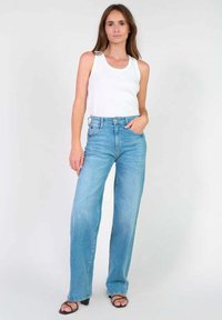 Jeans larges bleu clair en denim, à taille haute et légèrement délavés, associés à un top sans manches blanc et des sandales noires.