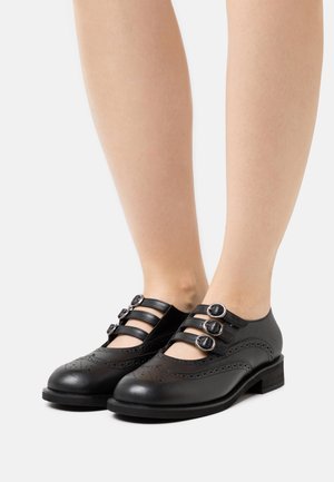 Chaussures Mary Jane en cuir noir avec trois brides à boucle et des détails brogues, portées pieds nus sur un fond blanc.