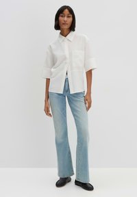 White oversized button-up shirt with short sleeves and a pocket, paired with light blue flared jeans and black flat shoes.