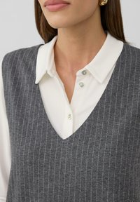 Vrouw die een witte colbertshirt draagt onder een grijze gestreepte mouwloze trui en gouden oorringen, gecropt tot aan de nek en schouders.