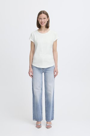 Camiseta blanca de manga corta con escote redondo, combinada con pantalones vaqueros de pierna ancha en azul claro y sandalias de tacón abierto y plateadas. Fondo liso.