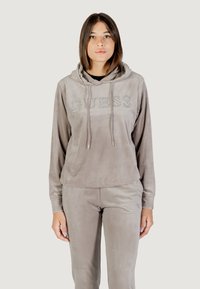 Sudadera de velour gris claro con "GUESS" en strass, que cuenta con un bolsillo tipo canguro en la parte frontal y capucha con cordón. Combinada con pantalones a juego.