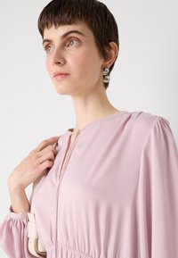 Vaaleanpunainen mekko, jossa on pyöreä pääntie, laskostetut hihat ja avainaukko-yksityiskohta. Mallilla on metalliset korvakorut ja hän pitää laukkuolkainta.