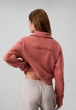 Kvinde med brunt hår iført cropped støvet lyserød Calvin Klein sweatshirt og lysegrå joggingbukser, stående med hånd på lænden.