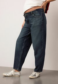 Blugi largi, de culoare albastru închis, confecționați din denim, cu un design cu cinci buzunare și cusături subtile. Asortați cu adidași argintii.