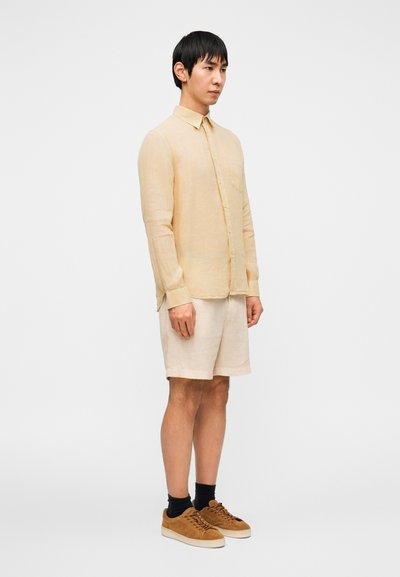 Ελαφρύ πουκάμισο από λινό κίτρινο χρώμα με μακριά μανίκια και γιακά με κουμπιά, σετ με μπεζ λινές βερμούδες και καφέ σουέτ αθλητικά παπούτσια.