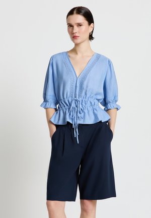 Femme debout les mains dans les poches, portant un chemisier bleu clair à col en V avec une taille à cordon et un short large bleu marine mi-genou.