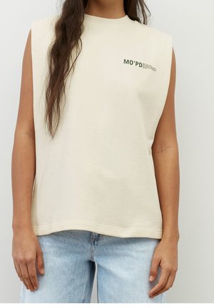 Femme portant un haut beige sans manches avec un petit texte noir "MO'PD" sur la poitrine gauche et un jean bleu clair sur fond uni.