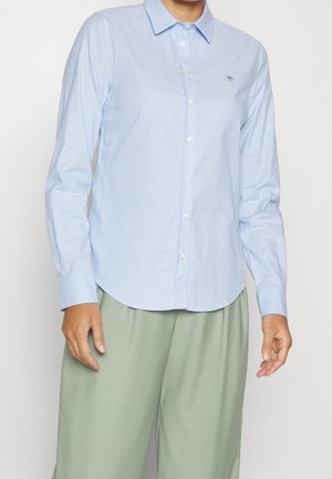 Osoba ubrana w jasnoniebieską koszulę zapinaną na guziki z ostrym kołnierzem oraz luźne jasnokrętkie spodnie na jednolitym tle.