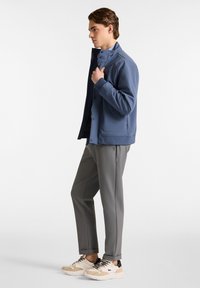 Blaue Softshell-Jacke mit hohem Kragen, kombiniert mit grauen Slim-Fit-Hosen und beigen Sneakern. Besitzt klare Linien und ein modernes Design.