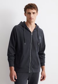Tmavě šedá mikina s kapucí na zip, která má přední kapsu, šňůrkovou kapuci a decentní logo na hrudi. Měkká, hladká tkanina s volným střihem.