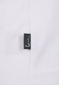 Balts audums ar gludu tekstūru, kas satur melnu apģērba etiķeti, kuri ir piešūta šuvē, ar minimālistisku baltu logotipu.