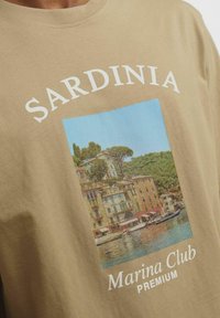 Beige kokvilnas T-krekls ar Vidusjūras ainavas attēlojumu, kurā redzami ēkas, ūdens un zaļās telpas. Uzraksts: "SARDINIA Marina Club PREMIUM."