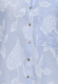 Chemise rayée bleu clair et blanc avec une grande broderie florale blanche et des boutons beige sur le devant, avec une poche poitrine.
