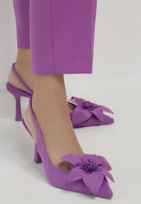 Tacchi alti slingback viola con punte affusolate e grandi decorazioni floreali in tessuto, indossati con pantaloni viola coordinati.