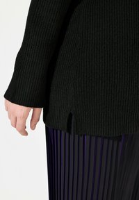 Schwarzer, gerippter Strickpullover mit einem Schlitz am Saum. Die Hand greift nach dem Stoff, der eine weiche Textur hat. Kombiniert mit plissierten lila Hosen.