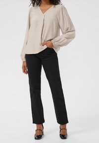 Blouse beige à manches longues avec un col en V, ornée d'un détail plissé, associée à un pantalon noir droit et des chaussures à talons noires.