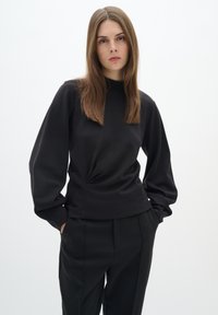 Black long-sleeve top with a high neck and gathered detail at the waist, paired with black trousers, showcasing a sleek, simple design.