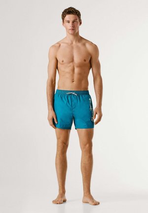 Junger Mann steht barfuß da, trägt türkise Badeshorts mit weißem Schriftlogo auf dem linken Bein vor einfarbigem Hintergrund.