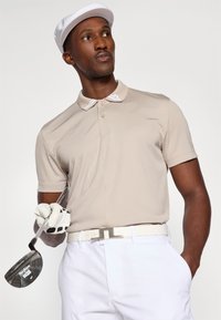 Beige polo skjorte med hvit krage, iført hvite bukser og et hvitt belte. Holder en golfkølle og har på seg en hvit caps.