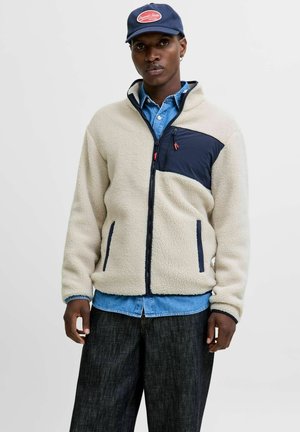 Fleece jacket in off-white with navy accents, zippered front, side pockets, and a chest pocket. Worn over a denim shirt with dark pants.