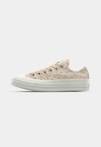 Sneakers sznurowane w jasnym beżu z teksturowanym wzorem sznurków, gumowym czubkiem i białą podeszwą. Posiadają niskoprofilowy kształt.