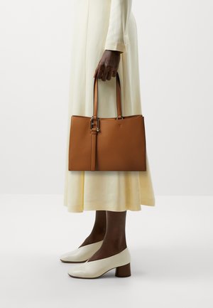 Borsa a tracolla in pelle color tan con doppi manici e un design elegante. Indossata con un vestito crema e scarpe con tacco bianche, con una finitura testurizzata.