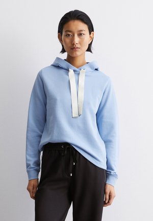 Helle blaue Kapuzenjacke aus weichem Material, mit einer Kordel mit weißen Akzenten. Getragen mit schwarzen Jogginghosen. Schlichte, lässige Passform.