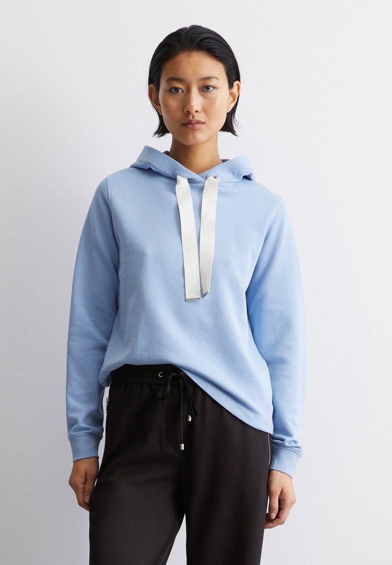 Helle blaue Kapuzenjacke aus weichem Material, mit einer Kordel mit weißen Akzenten. Getragen mit schwarzen Jogginghosen. Schlichte, lässige Passform.
