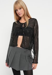 Jaqueta cropped preta com lantejoulas, mangas longas, fecho frontal tipo laço e textura transparente, combinada com uma saia mini plissada cinza.