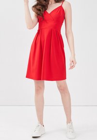 Robe rouge sans manches avec un décolleté en V, un corsage ajusté et une jupe évasée. Tissu lisse et plis subtils. Portée avec des baskets blanches.